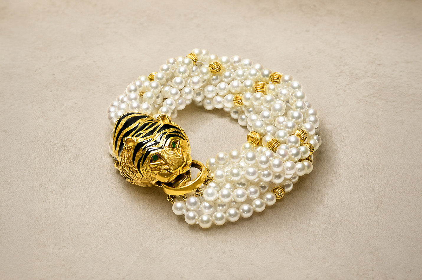 Multi-strand pearl bracelet with gold beads and a tiger head clasp in yellow gold, black enamel, and green eyes on a soft beige background.