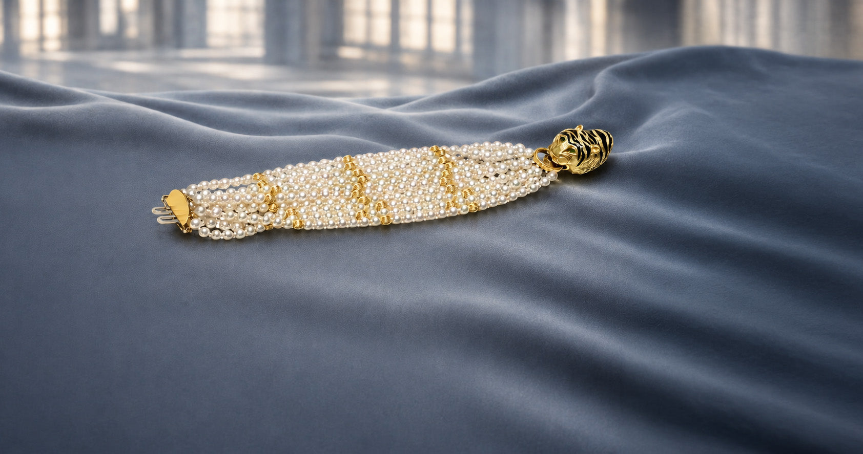 Multi-strand pearl bracelet with gold bead accents and a black enamel tiger-head clasp displayed on dusty blue-gray satin.